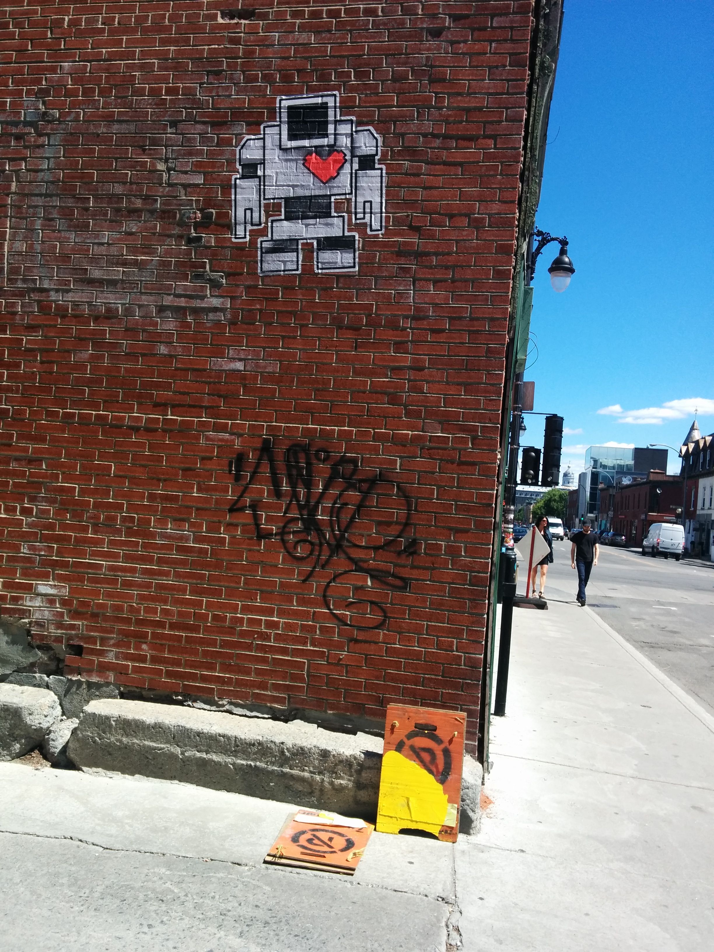 There`s a graffiti tag on the corner of an alley way. Above is is a white robot wheat pase with a red hard.